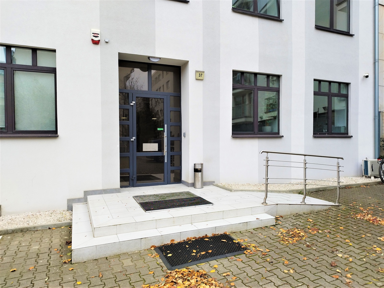 Fotografia przedstawia wejście do budynku, w którym na 2 piętrze zlokalizowane jest biuro Stowarzyszenia Metropolia Poznań. Znajduje się ono od strony dziedzińca parkingowego i wyposażone zostało w rampę najazdową w celu umożliwienia dostępności obiektu dla osób z niepełnosprawnością ruchu, ze względu na widoczne występowanie schodów dwustopniowych. Drzwi ukazane na fotografii otworzyć można jedynie za pomocą kodu, który wpisywany jest poprzez użycie czytnika zamontowanego na elewacji budynku na wysokości ok. 100 cm.